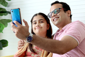 Wall Mural - Indian happy family spending time together, cute daughter girl kid in India traditional dress costume and her father taking a selfie by mobile phone while sitting in living room or making video call.