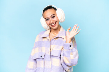 Canvas Print - Young Arab woman wearing winter muffs isolated on blue background saluting with hand with happy expression