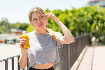 Wall Mural - Young blonde woman holding an orange juice at outdoors having doubts and with confuse face expression