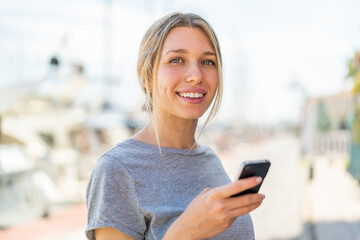 Wall Mural - Young blonde woman using mobile phone at outdoors smiling a lot