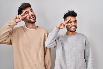 Wall Mural - Young homosexual couple standing over white background doing peace symbol with fingers over face, smiling cheerful showing victory