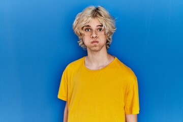 Sticker - Young modern man standing over blue background puffing cheeks with funny face. mouth inflated with air, crazy expression.