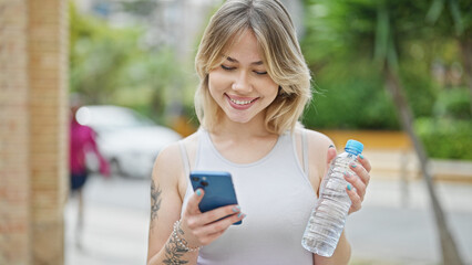 Canvas Print - Young blonde woman using smartphone holding bottle of water at street