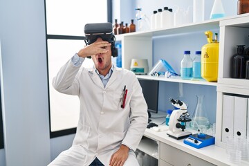 Sticker - Young hispanic man working at scientist laboratory wearing vr glasses peeking in shock covering face and eyes with hand, looking through fingers afraid