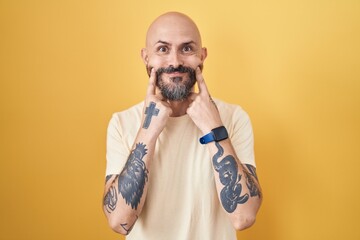 Sticker - Hispanic man with tattoos standing over yellow background smiling with open mouth, fingers pointing and forcing cheerful smile