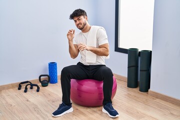 Poster - Hispanic man with beard sitting on pilate balls at yoga room disgusted expression, displeased and fearful doing disgust face because aversion reaction. with hands raised