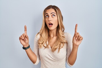 Poster - Young blonde woman standing over isolated background amazed and surprised looking up and pointing with fingers and raised arms.