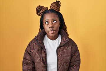 African woman with braided hair standing over yellow background making fish face with lips, crazy and comical gesture. funny expression.