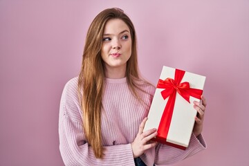 Sticker - Young caucasian woman holding gift smiling looking to the side and staring away thinking.