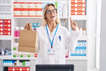 Sticker - Young blonde woman working at pharmacy drugstore holding paper bag surprised with an idea or question pointing finger with happy face, number one