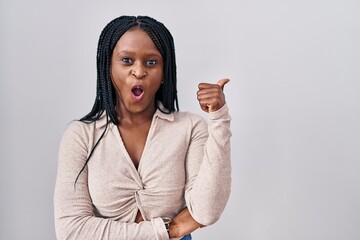 Sticker - African woman with braids standing over white background surprised pointing with hand finger to the side, open mouth amazed expression.