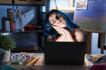 Canvas Print - Young modern girl with blue hair sitting at art studio with laptop at night sleeping tired dreaming and posing with hands together while smiling with closed eyes.