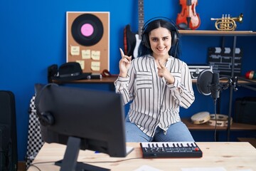 Poster - Young modern girl with blue hair at music studio wearing headphones smiling and looking at the camera pointing with two hands and fingers to the side.