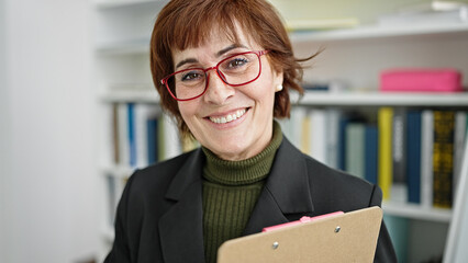 Sticker - Mature hispanic woman teacher standing by white board with clipboard at library university