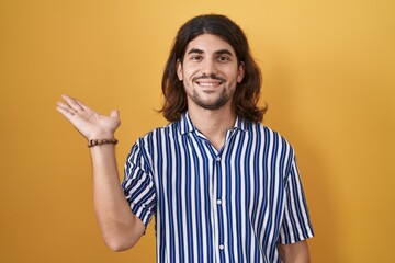 Sticker - Hispanic man with long hair standing over yellow background smiling cheerful presenting and pointing with palm of hand looking at the camera.