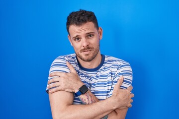 Sticker - Young hispanic man standing over blue background shaking and freezing for winter cold with sad and shock expression on face