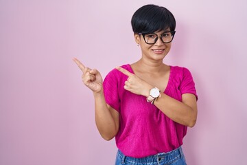 Poster - Young asian woman with short hair standing over pink background smiling and looking at the camera pointing with two hands and fingers to the side.