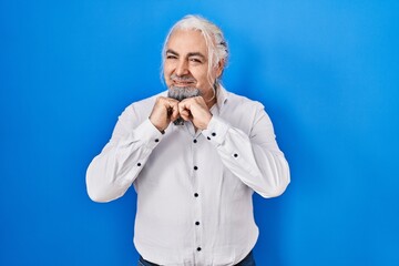 Wall Mural - Middle age man with grey hair standing over blue background laughing nervous and excited with hands on chin looking to the side