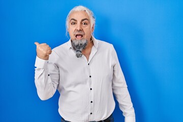 Sticker - Middle age man with grey hair standing over blue background surprised pointing with hand finger to the side, open mouth amazed expression.