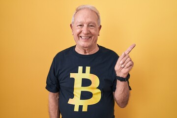 Wall Mural - Senior man with grey hair wearing bitcoin t shirt with a big smile on face, pointing with hand finger to the side looking at the camera.