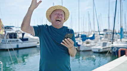 Senior grey-haired man tourist wearing summer hat using smartphone with winner expression at port