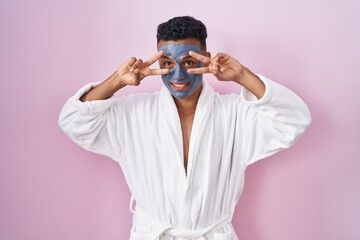 Sticker - Young hispanic man wearing beauty face mask and bath robe doing peace symbol with fingers over face, smiling cheerful showing victory