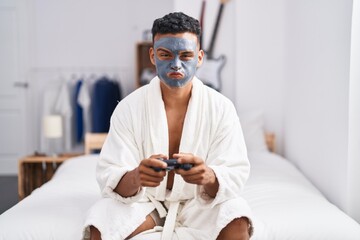 Canvas Print - Young hispanic man wearing beauty face mask playing video games puffing cheeks with funny face. mouth inflated with air, catching air.