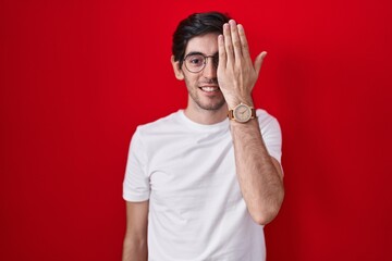 Wall Mural - Young hispanic man standing over red background covering one eye with hand, confident smile on face and surprise emotion.