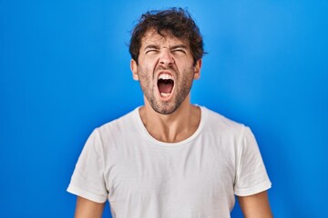 Sticker - Hispanic young man standing over blue background angry and mad screaming frustrated and furious, shouting with anger. rage and aggressive concept.