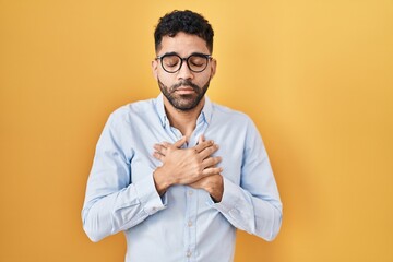 Wall Mural - Hispanic man with beard standing over yellow background smiling with hands on chest with closed eyes and grateful gesture on face. health concept.