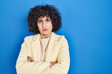 Poster - Young brunette woman with curly hair standing over blue background skeptic and nervous, disapproving expression on face with crossed arms. negative person.