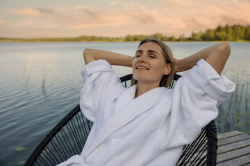 Wall Mural - woman in white bathrobe relaxing in chair on lake footbridge at warm summer evening. nature enjoyment