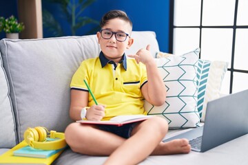 Poster - Young hispanic kid doing homework sitting on the sofa smiling doing phone gesture with hand and fingers like talking on the telephone. communicating concepts.