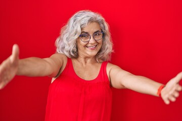 Wall Mural - Middle age woman with grey hair standing over red background looking at the camera smiling with open arms for hug. cheerful expression embracing happiness.