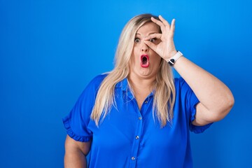 Canvas Print - Caucasian plus size woman standing over blue background doing ok gesture shocked with surprised face, eye looking through fingers. unbelieving expression.