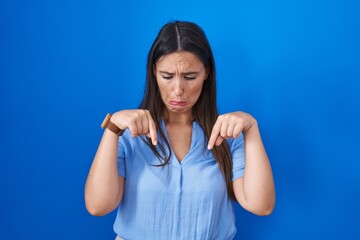 Canvas Print - Young brunette woman standing over blue background pointing down looking sad and upset, indicating direction with fingers, unhappy and depressed.