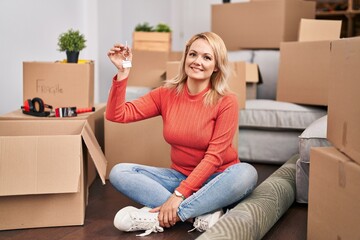 Canvas Print - Blonde woman holding keys of new home sitting on the floor looking positive and happy standing and smiling with a confident smile showing teeth