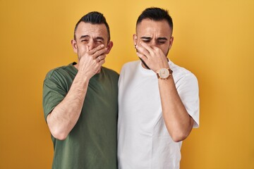 Sticker - Homosexual couple standing over yellow background smelling something stinky and disgusting, intolerable smell, holding breath with fingers on nose. bad smell