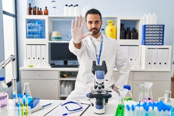 Sticker - Young hispanic man with beard working at scientist laboratory doing stop sing with palm of the hand. warning expression with negative and serious gesture on the face.