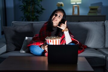 Canvas Print - Hispanic woman eating popcorn watching a movie on the sofa smiling doing phone gesture with hand and fingers like talking on the telephone. communicating concepts.