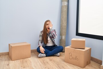 Wall Mural - Young blonde woman sitting on the floor moving to a new home covering one eye with hand, confident smile on face and surprise emotion.