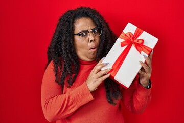 Poster - Plus size hispanic woman holding gift in shock face, looking skeptical and sarcastic, surprised with open mouth