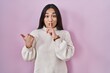 Young south asian woman standing over pink background asking to be quiet with finger on lips pointing with hand to the side. silence and secret concept.