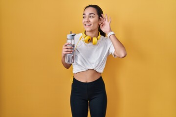 Wall Mural - Young south asian woman wearing sportswear drinking water smiling with hand over ear listening an hearing to rumor or gossip. deafness concept.