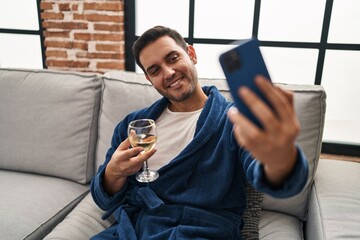 Wall Mural - Young hispanic man make selfie by smartphone drinking wine at home