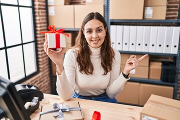 Sticker - Young hispanic doctor woman working at small business ecommerce holding present smiling happy pointing with hand and finger to the side