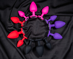 A set of silicone anal plugs in different colors and sizes on a black silk sheet. 