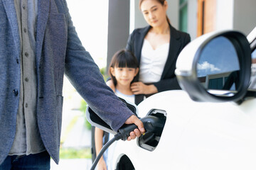 Focus closeup hand charging electric car, insert charger device into electric vehicle as progressive lifestyle concept of alternative green energy technology with blurred family in the background.