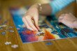 © MNStudio - Close-up on woman hand playing puzzles at home. Connecting jigsaw puzzle pieces in a living room table, assembling a jigsaw puzzle.