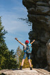 © junky_jess - Woman hiker with backpack and bandana standing on the rocks with raising hands among the tree tops in summer day, vertical photo, selected focus.